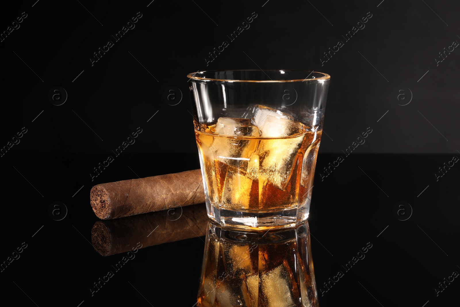 Photo of Glass of whiskey with ice cubes and cigar on black mirror surface