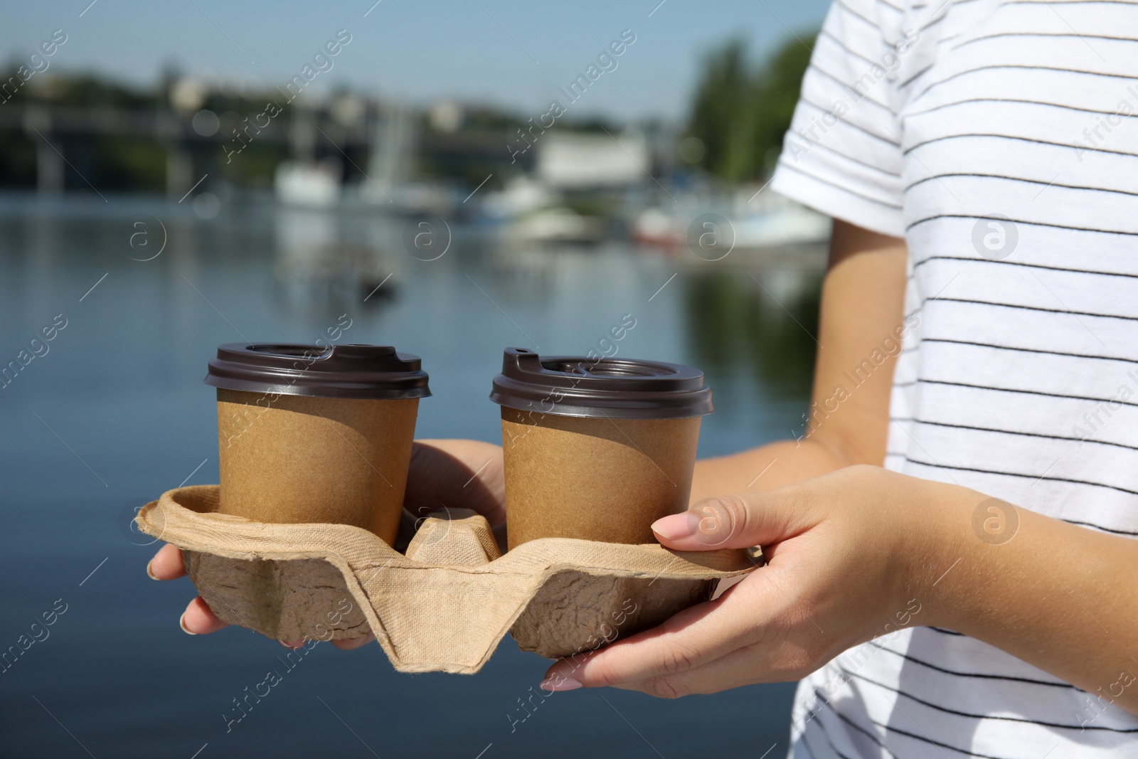 Photo of Woman holding takeaway paper coffee cups with plastic lids in cardboard holder near river, closeup