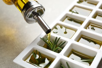 Photo of Pouring olive oil into ice cube tray with fresh rosemary at grey table, closeup