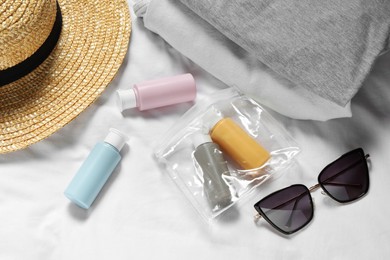 Photo of Cosmetic travel kit. Plastic bag with small containers of personal care products, beach accessories and clothes on bed, above view