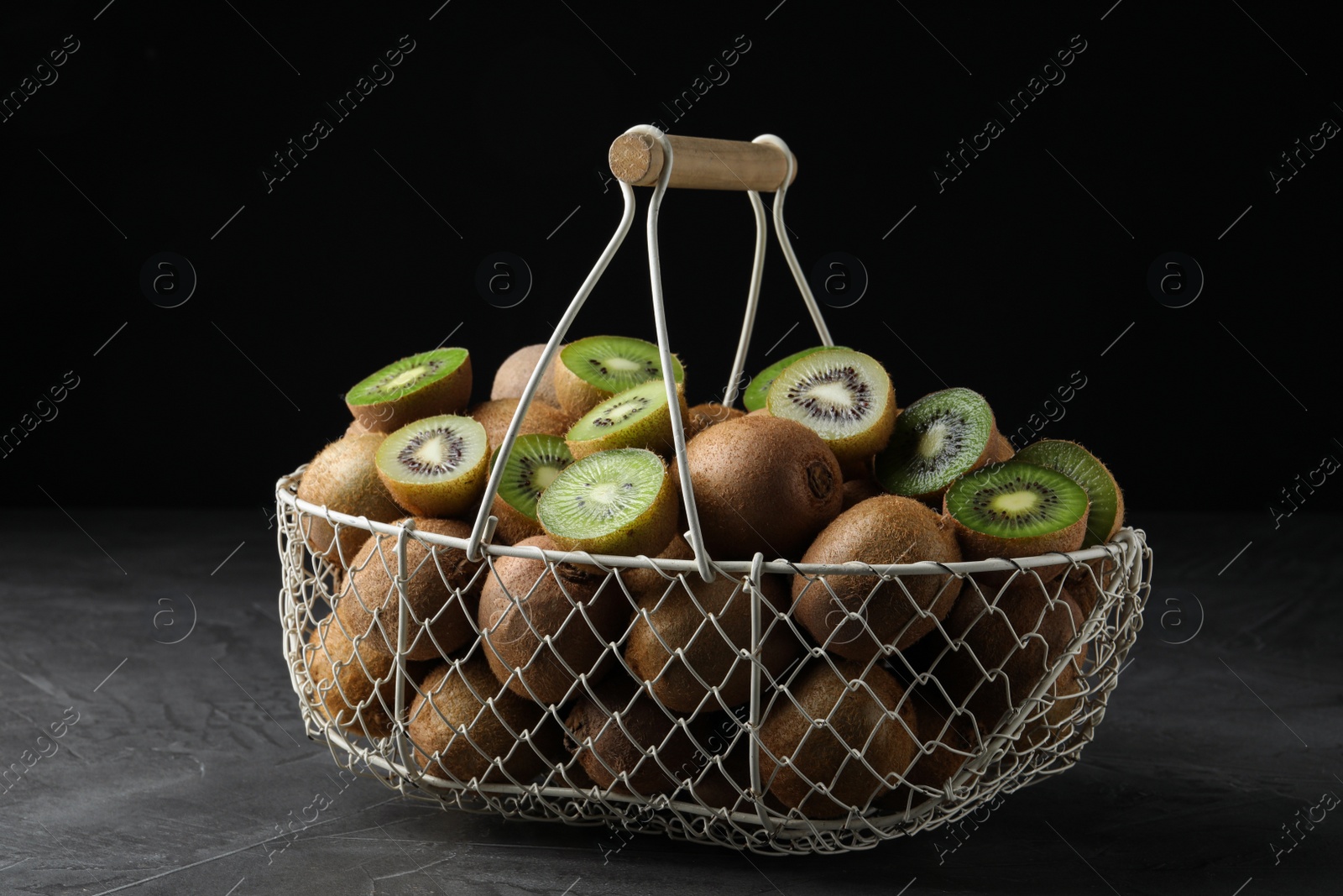 Photo of Fresh ripe kiwis in metal basket on grey table against dark background