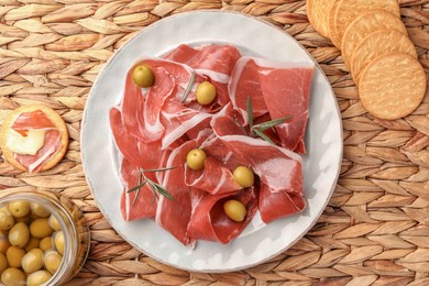 Photo of Slices of tasty cured ham, olives and rosemary on wicker mat, flat lay