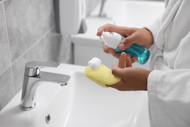 Photo of Young man applying face cleanser on sponge above sink in bathroom, closeup