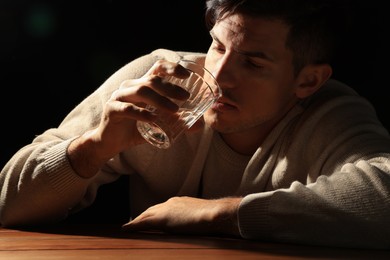 Photo of Addicted man drinking alcohol at wooden table against black background