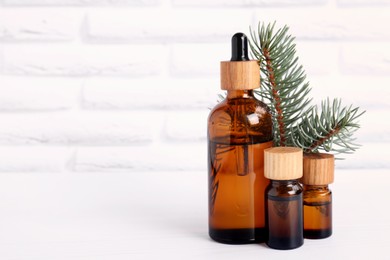 Bottles of essential oil and pine branch on white wooden table near brick wall, space for text