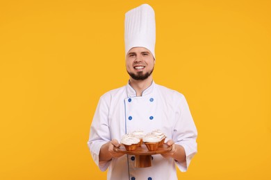 Photo of Happy professional confectioner in uniform holding delicious cupcakes on yellow background