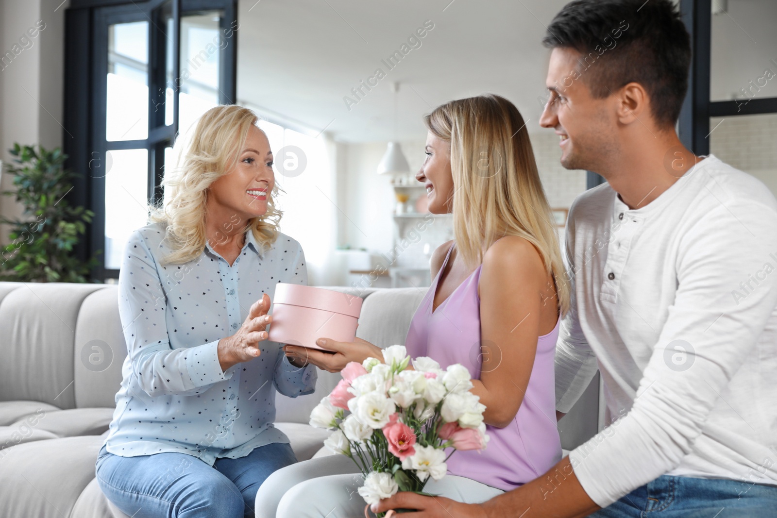 Photo of Young couple with bouquet and gift box congratulating their mother in living room