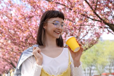 Photo of Beautiful young woman holding paper coffee cup in park with blossoming sakura trees