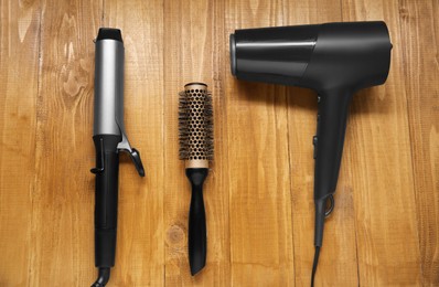 Photo of Curling iron, round brush and hair dryer on wooden background, flat lay