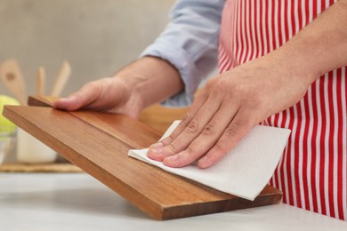 Photo of Man wiping wooden cutting board with paper napkin at white table, closeup