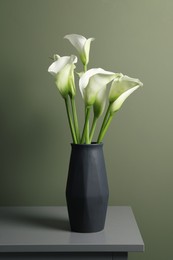 Beautiful calla lily flowers in vase on grey table near olive wall