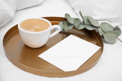Photo of Tray with cup of coffee, envelope and eucalyptus branch on white bed, closeup