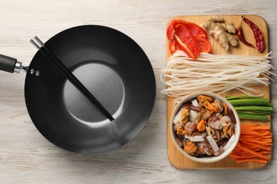 Photo of Black wok, chopsticks and board with products on light wooden table, top view