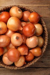 Wicker basket with many ripe onions on wooden table, top view