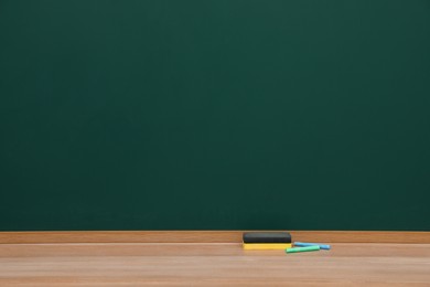 Photo of Colorful chalks and duster on wooden table near green board. Space for text