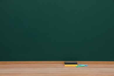 Colorful chalks and duster on wooden table near green board. Space for text