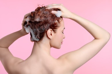 Young woman washing her hair with shampoo on pink background, back view