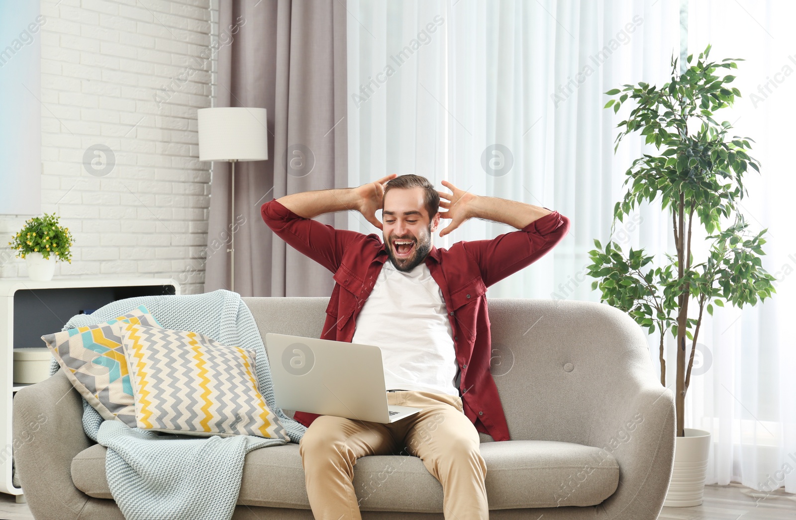 Photo of Emotional young man with laptop celebrating victory on sofa at home