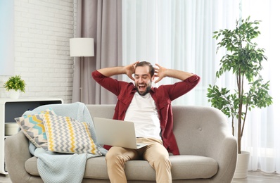 Emotional young man with laptop celebrating victory on sofa at home