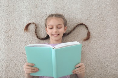 Cute little girl reading book on floor at home, top view