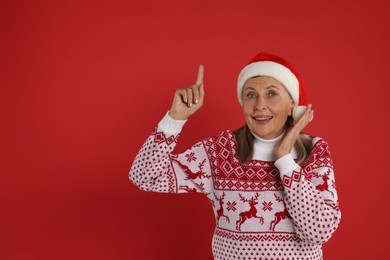 Photo of Happy senior woman in Christmas sweater and Santa hat pointing at something on red background. Space for text
