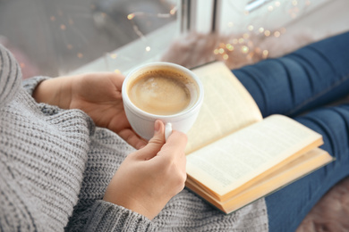 Woman with cup of coffee reading book at home, closeup