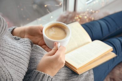 Photo of Woman with cup of coffee reading book at home, closeup