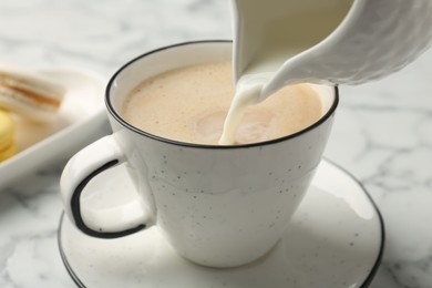 Photo of Pouring milk from pitcher into cup with coffee at white marble table, closeup