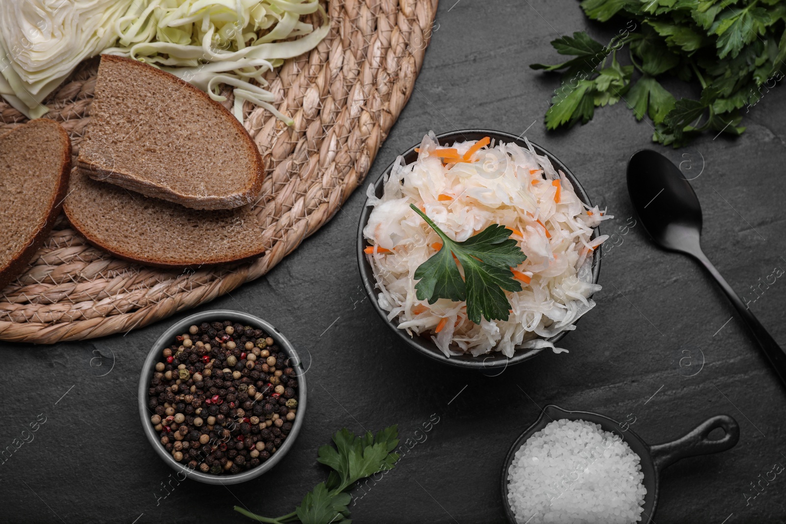 Photo of Bowl of tasty sauerkraut and ingredients on black table, flat lay