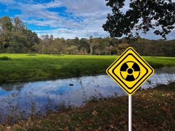 Image of Radioactive pollution. Yellow warning sign with hazard symbol near river outdoors