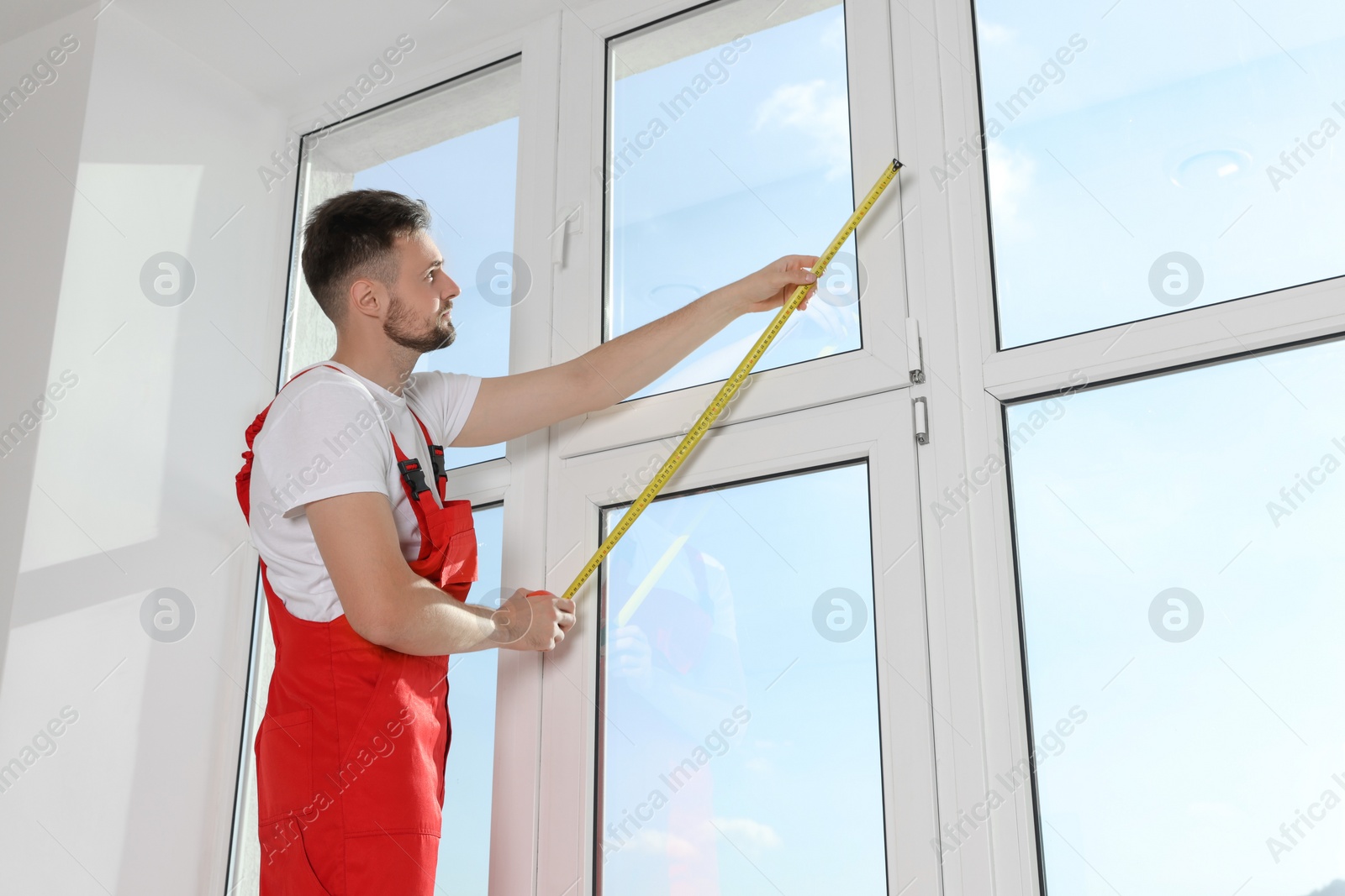 Photo of Worker in uniform measuring window with tape indoors. Roller blinds installation