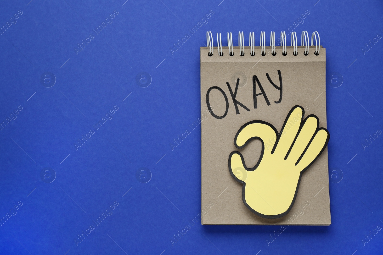 Photo of Notepad with word Okay and paper cutout of OK hand gesture on blue background, top view. Space for text