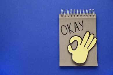 Photo of Notepad with word Okay and paper cutout of OK hand gesture on blue background, top view. Space for text