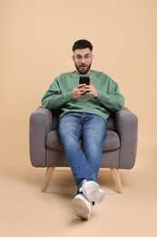 Emotional young man using smartphone in armchair on beige background