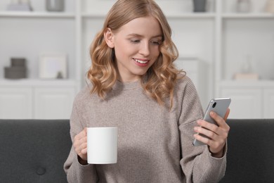 Photo of Beautiful woman with blonde hair holding cup and smartphone on sofa indoors