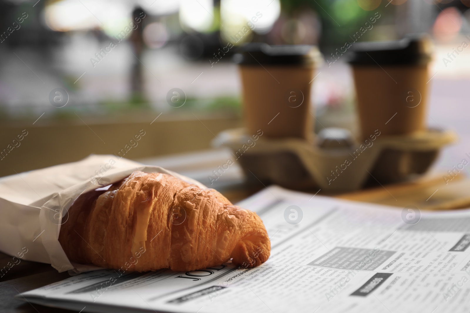 Photo of Tasty croissant, newspaper and paper cups of coffee on wooden table outdoors