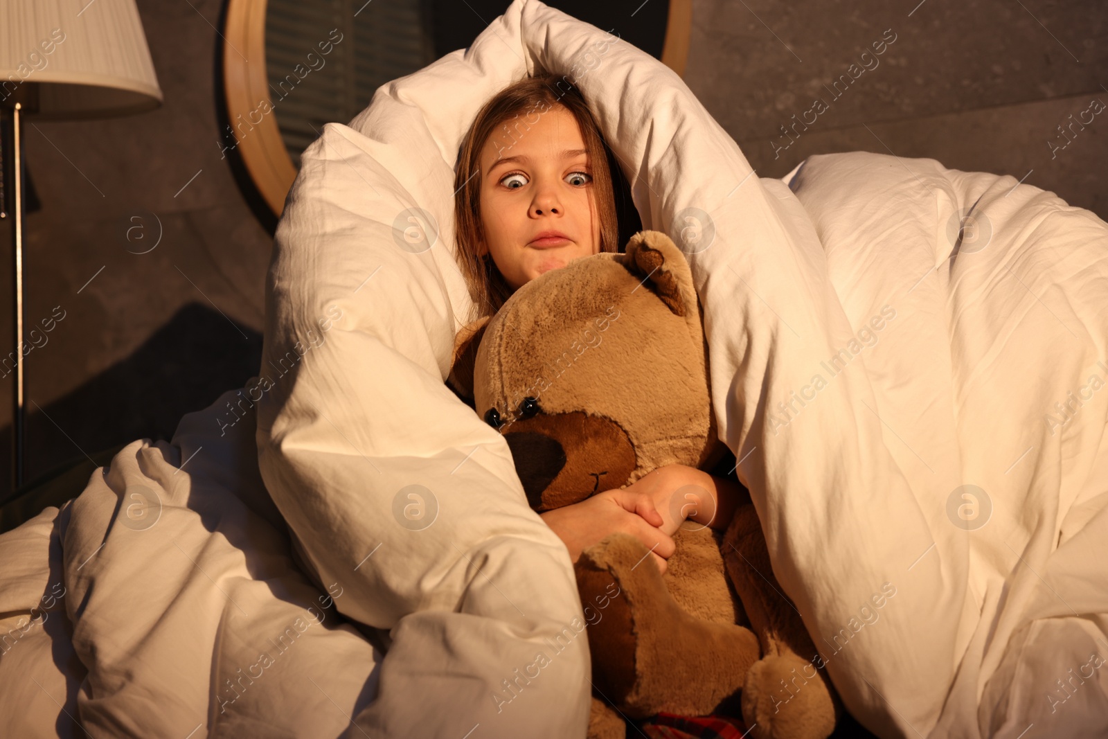 Photo of Scared little girl with toy hiding from monster in duvet at night