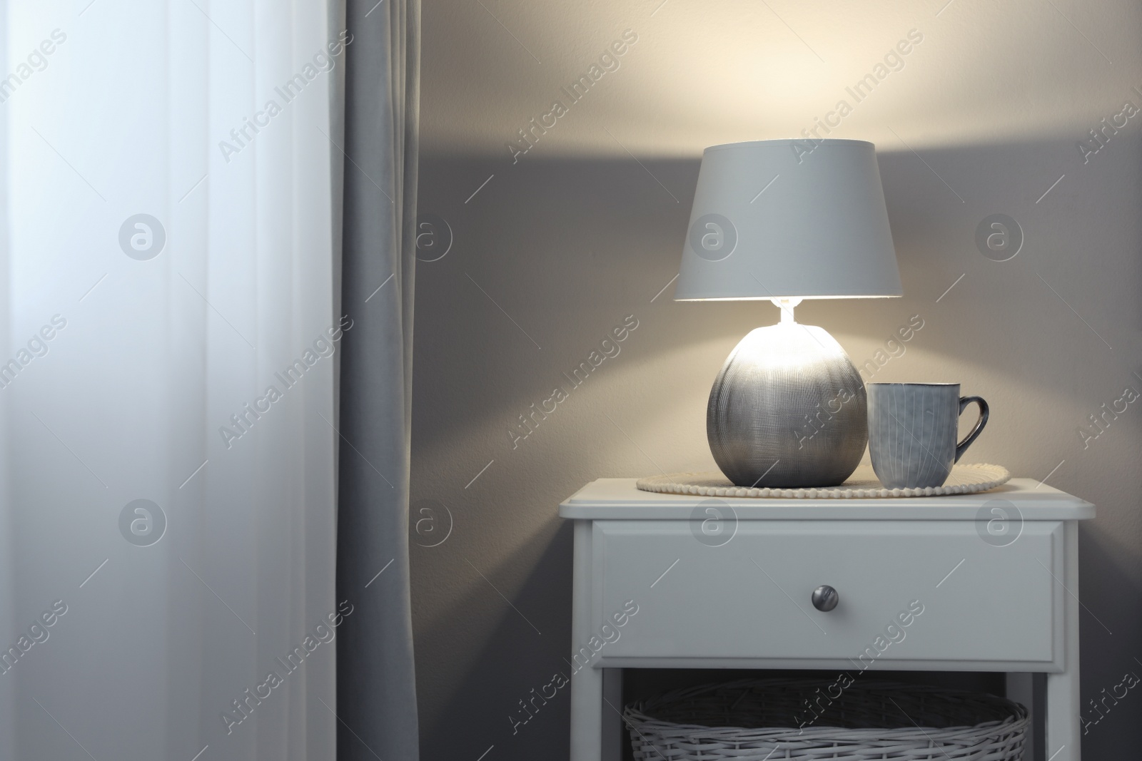 Photo of Stylish lamp and cup of drink on white nightstand in room, space for text