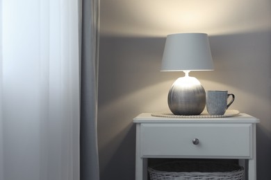 Stylish lamp and cup of drink on white nightstand in room, space for text