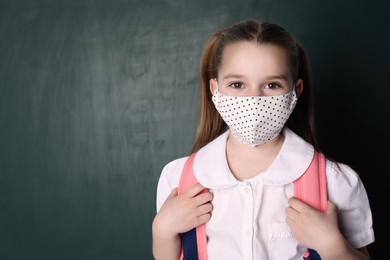 Little girl wearing protective mask and backpack near chalkboard, space for text. Child safety