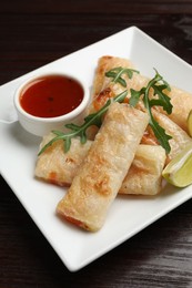 Photo of Tasty fried spring rolls, arugula, lime and sauce on wooden table, closeup