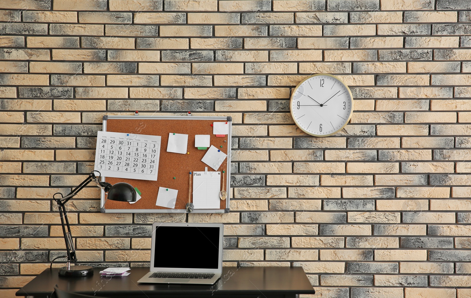 Photo of Stylish workplace with laptop, cork board and clock on brick wall. Time management