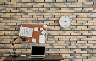 Stylish workplace with laptop, cork board and clock on brick wall. Time management