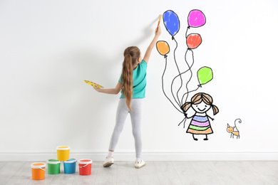 Image of Cute child drawing girl with balloons and cat on white wall indoors