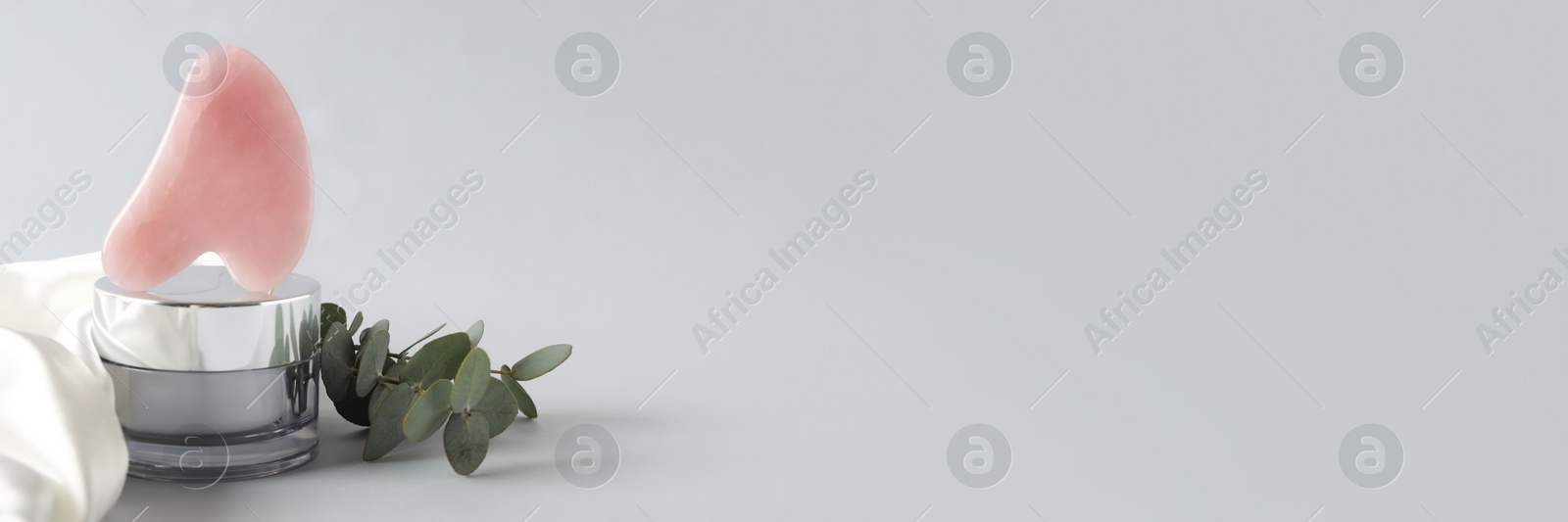 Image of Rose quartz gua sha tool, jar of cream and eucalyptus branches on grey background, space for text. Banner design