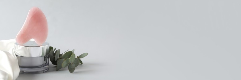 Image of Rose quartz gua sha tool, jar of cream and eucalyptus branches on grey background, space for text. Banner design