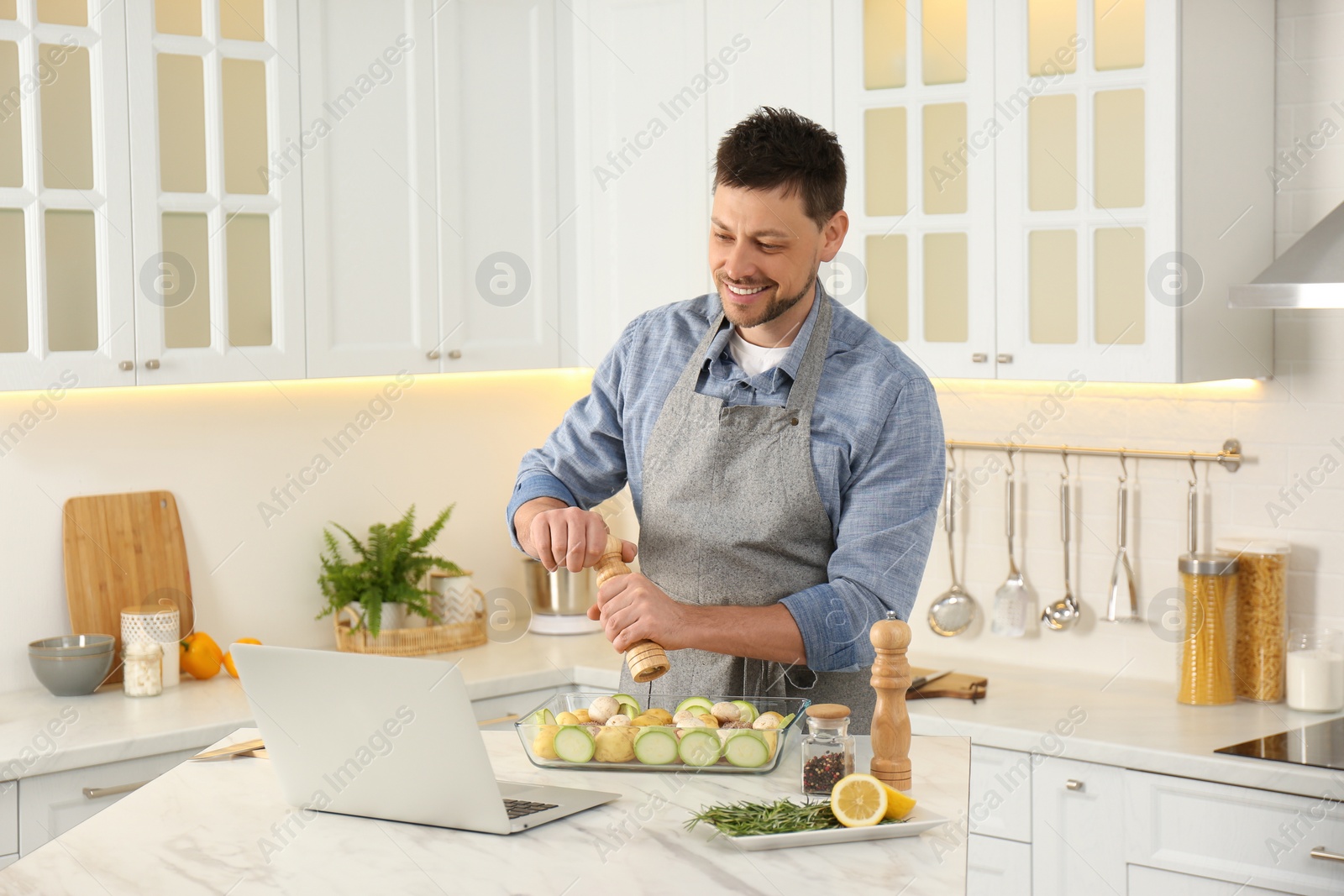 Photo of Man adding spices to dish while watching online cooking course via laptop in kitchen