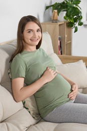 Photo of Happy pregnant woman sitting on sofa and touching her belly in living room