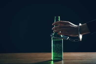 Photo of Alcohol addiction. Woman handcuffed to bottle of beer against black background, closeup. Space for text