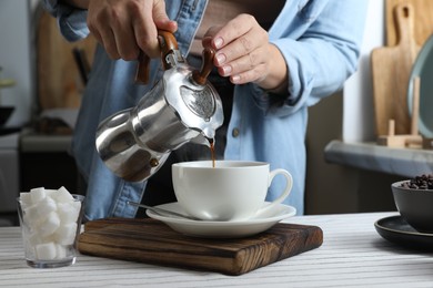 Woman pouring aromatic coffee from moka pot into cup at table in kitchen, closeup